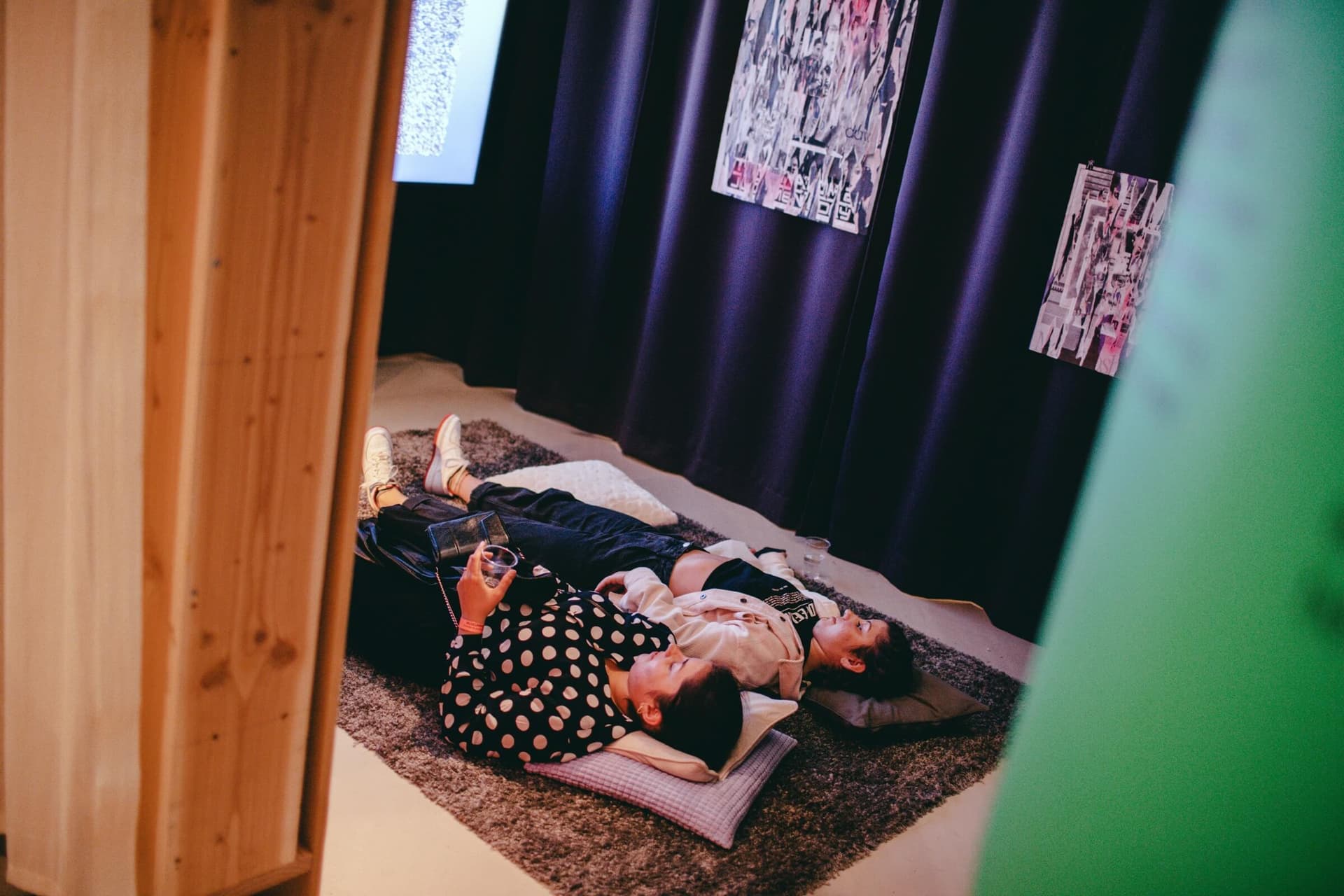 Two people are lying on a carpet with cushions under their heads in a room in the exhibition Designing the Social. They are looking at a screen, of which only a small part is visible in the photograph. The room is separated on one side by… 