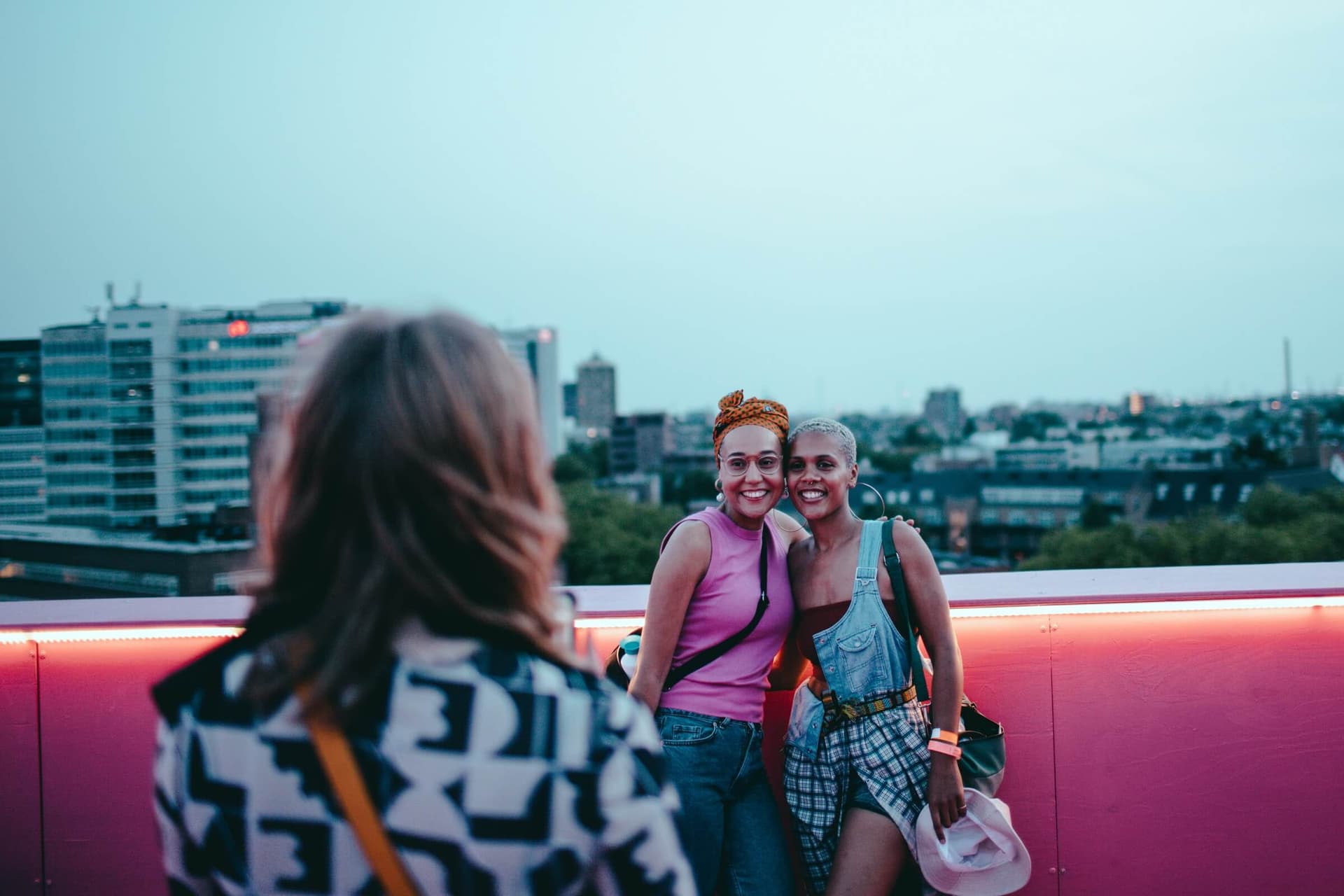 The pink Stage at dusk. A person with medium-length blond hair and a blouse or jacket with a black and white motif stands with his back to the viewer. She seems to be taking a photograph of two other women who have put their arms around ea… 