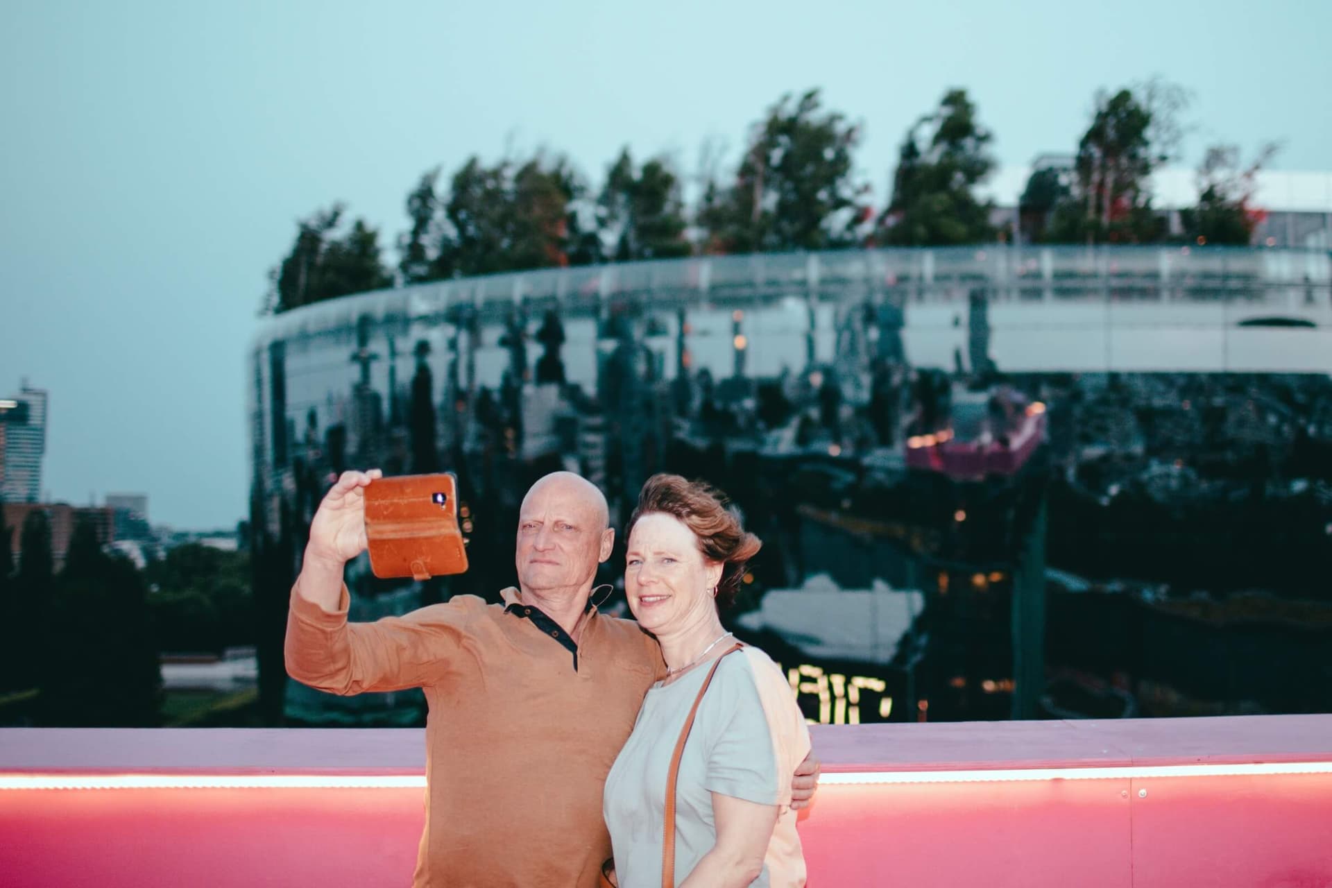 The pink Stage at dusk. A man and a woman are standing next to each other. The man has his arm around the woman. In his free hand, he holds his phone to take a selfie of the two of them. The Depot can be seen in the background.  