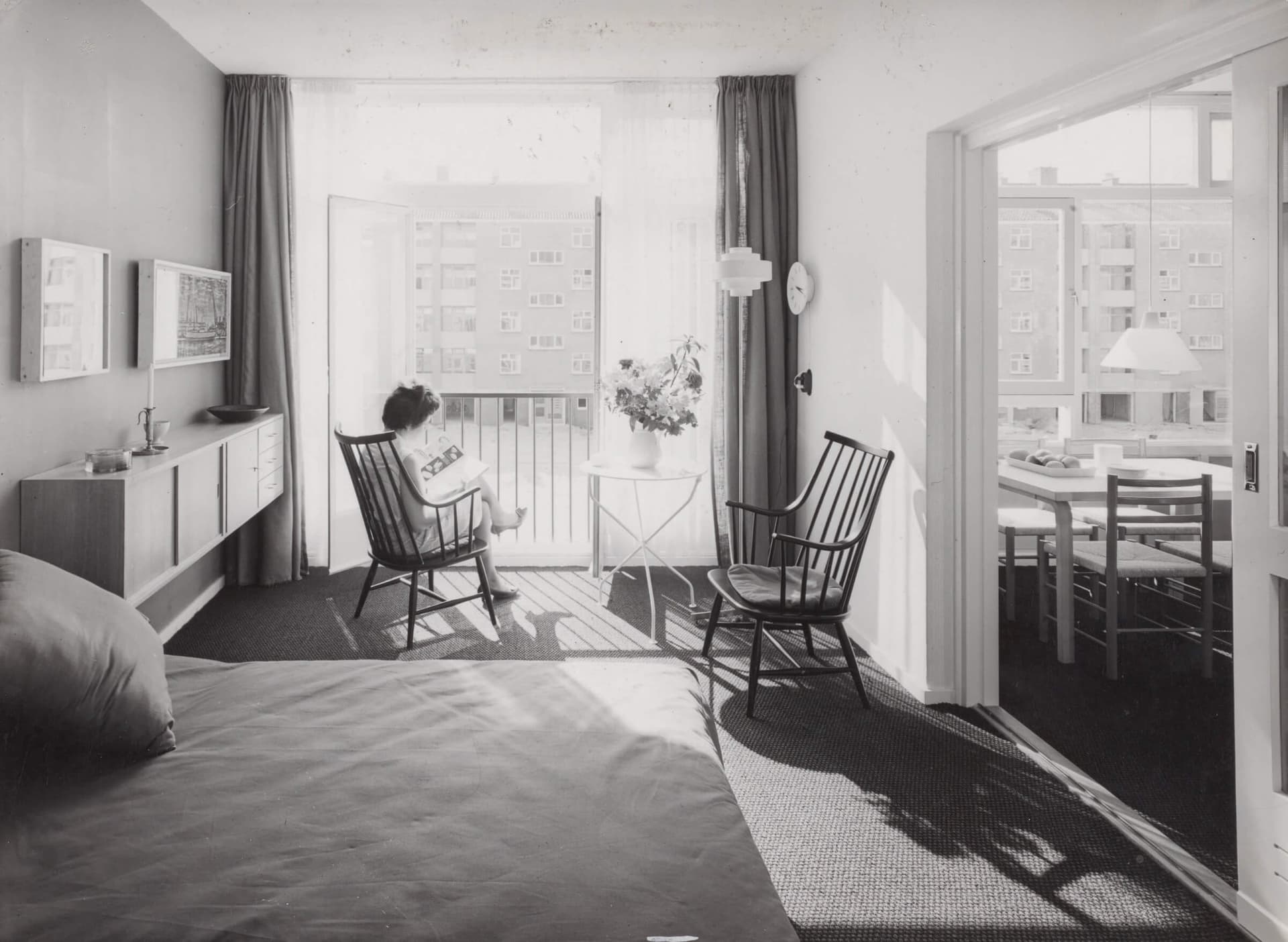 Zwartwitfoto van een ingerichte modelwoning. Iemand zit op een stoel voor een groot raam.
