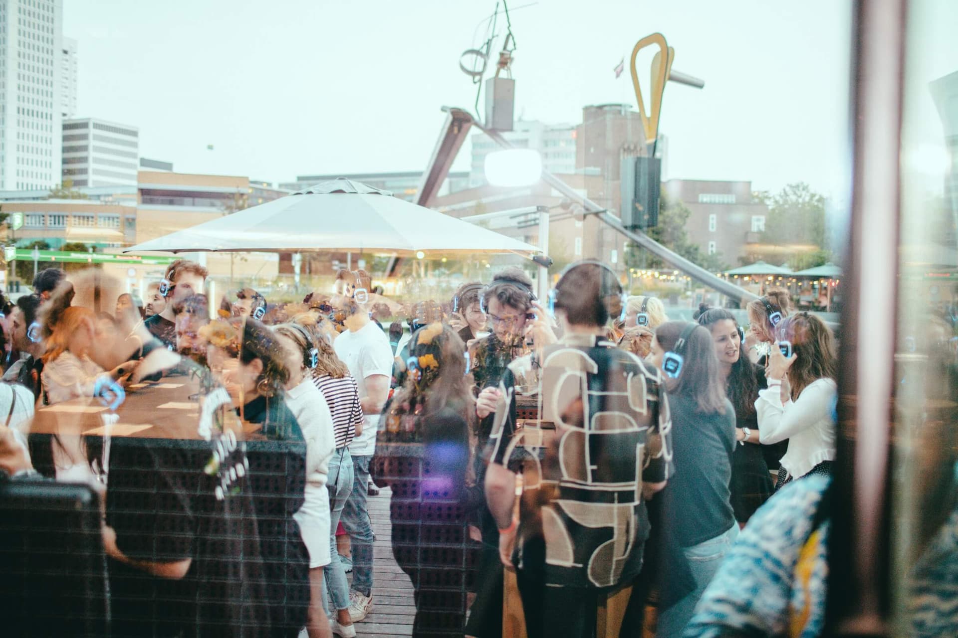 A group of people is visible through a reflected glass window. They are wearing headphones with blue light. They are standing on the terrace of Het Nieuwe Instituut. Behind this group, a brown parasol and the work of art by Auke de Vries i… 