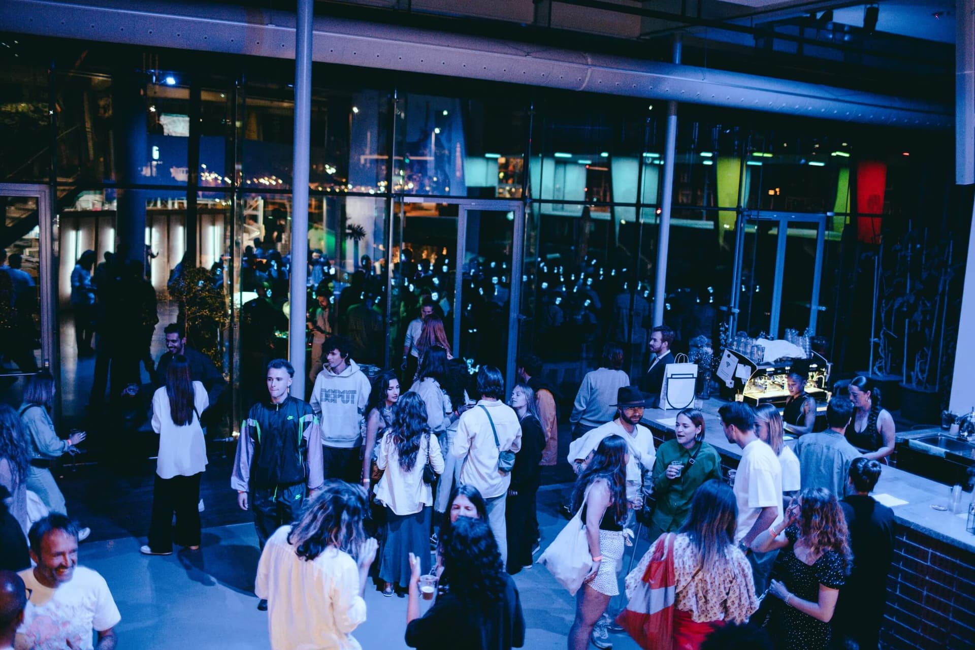 The New Café photographed from above. Behind the bar, two people are working. The space is further filled with people standing in groups and talking to each other and/or looking around. Through the glass windows of the café, the people on… 