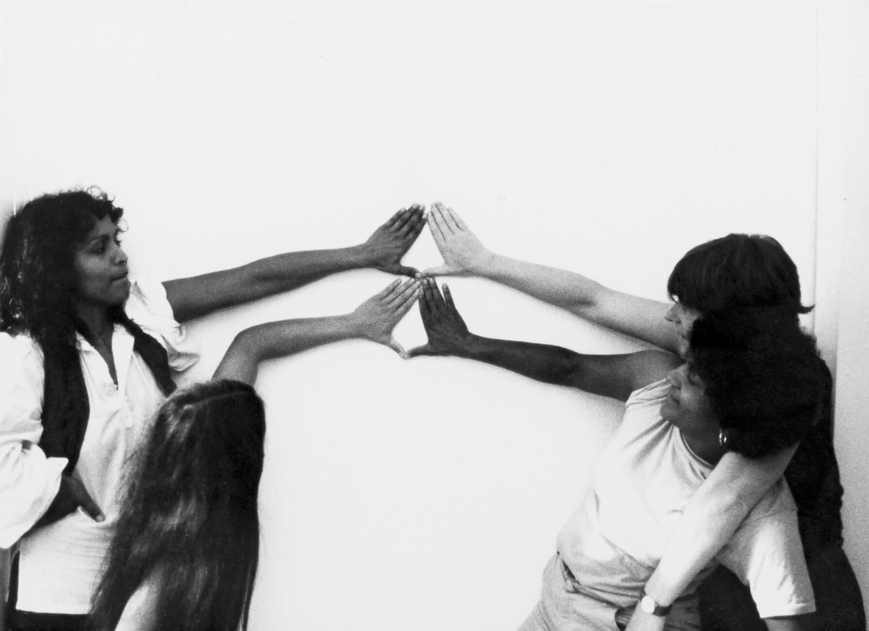 Vier vrouwen vormen driehoeken of het Yoni-teken met hun handen tegen een witte muur. Tegen de klok in vanaf rechtsonder: Tania Leon, Lida van den Broek, Ananda Spies, Margrethe Rumeser. Jaar: 198?. Foto: Gon Buurman. Bron: Collectie IAV-Atria   