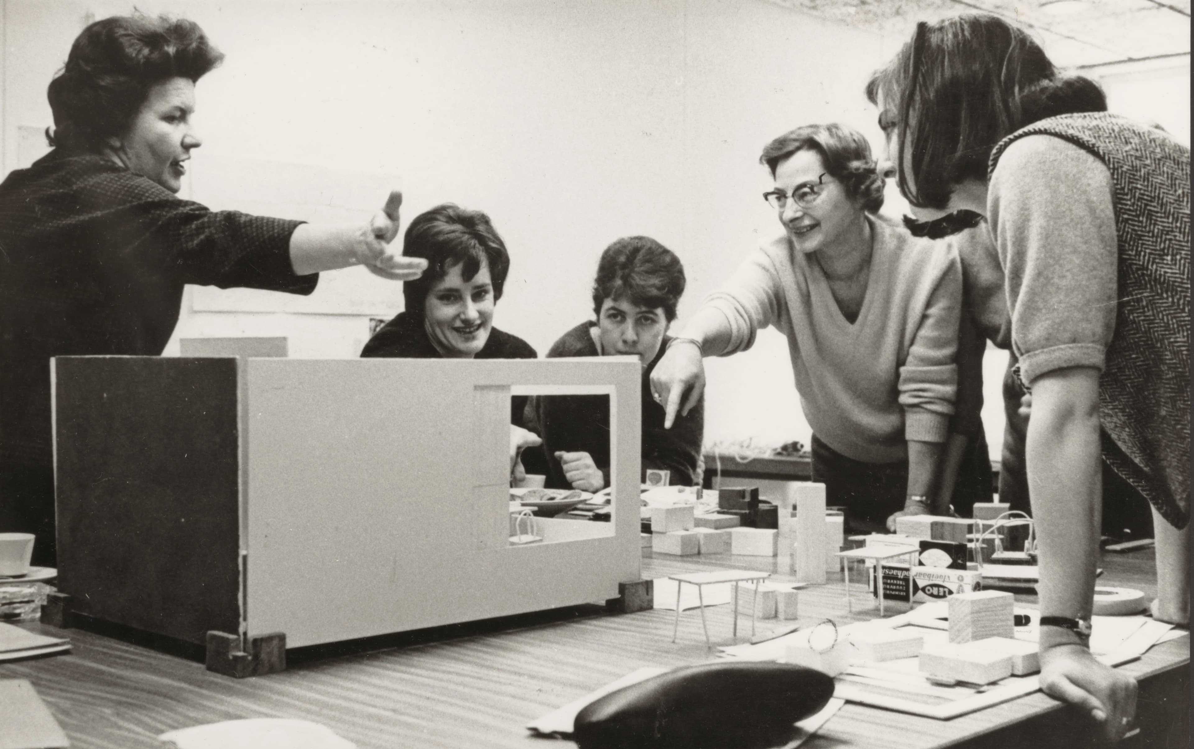 A group of women are located around a table, three of them are standing, two of those women are pointing to a building model. The remaining two women are sitting and are looking at it as well.