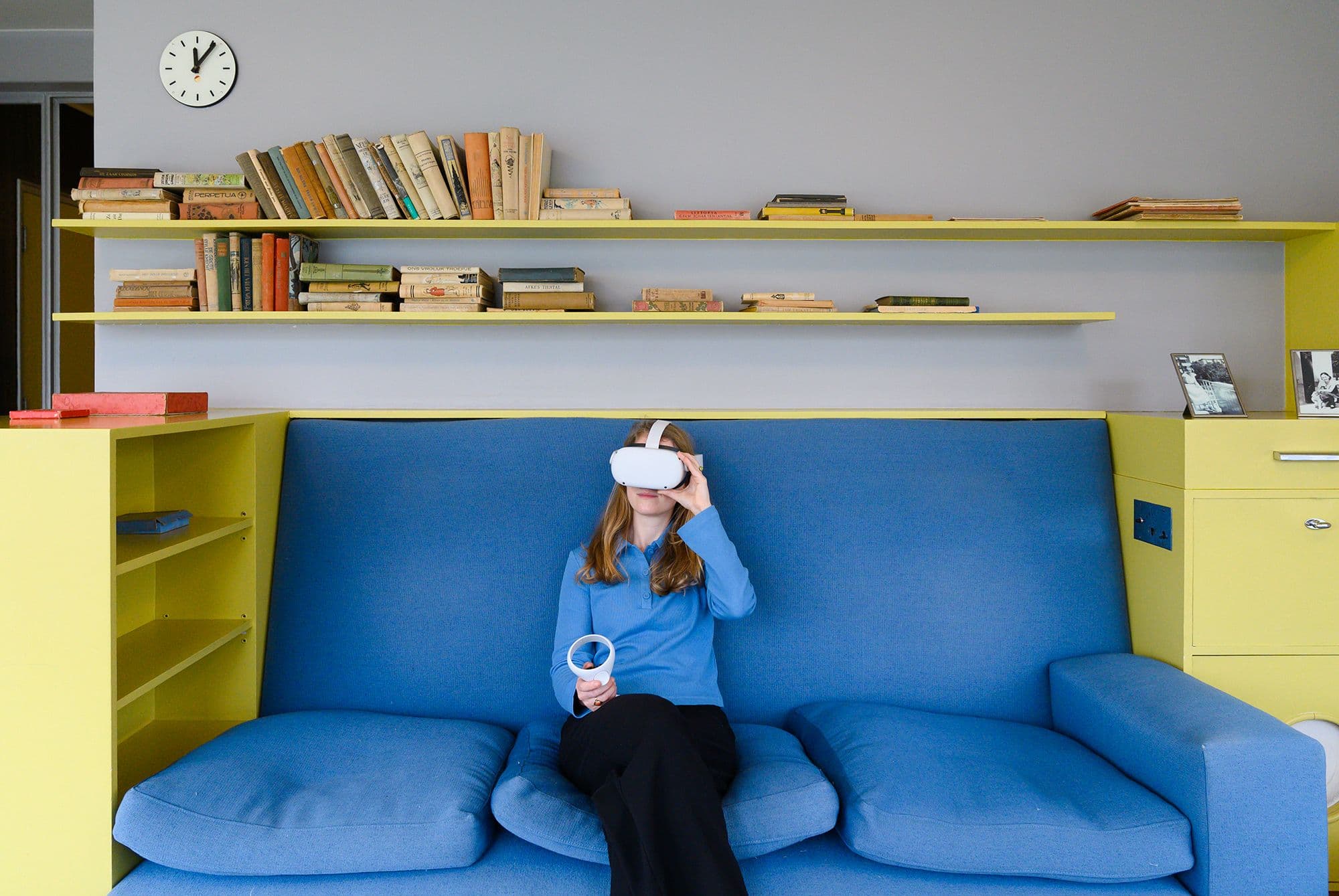 Een vrouw met een virtualreality-headset zit op een blauwe bank. Achter haar zijn gele planken vol boeken. De ruimte heeft een moderne uitstraling met contrasterende kleuren en een retro-accent.
