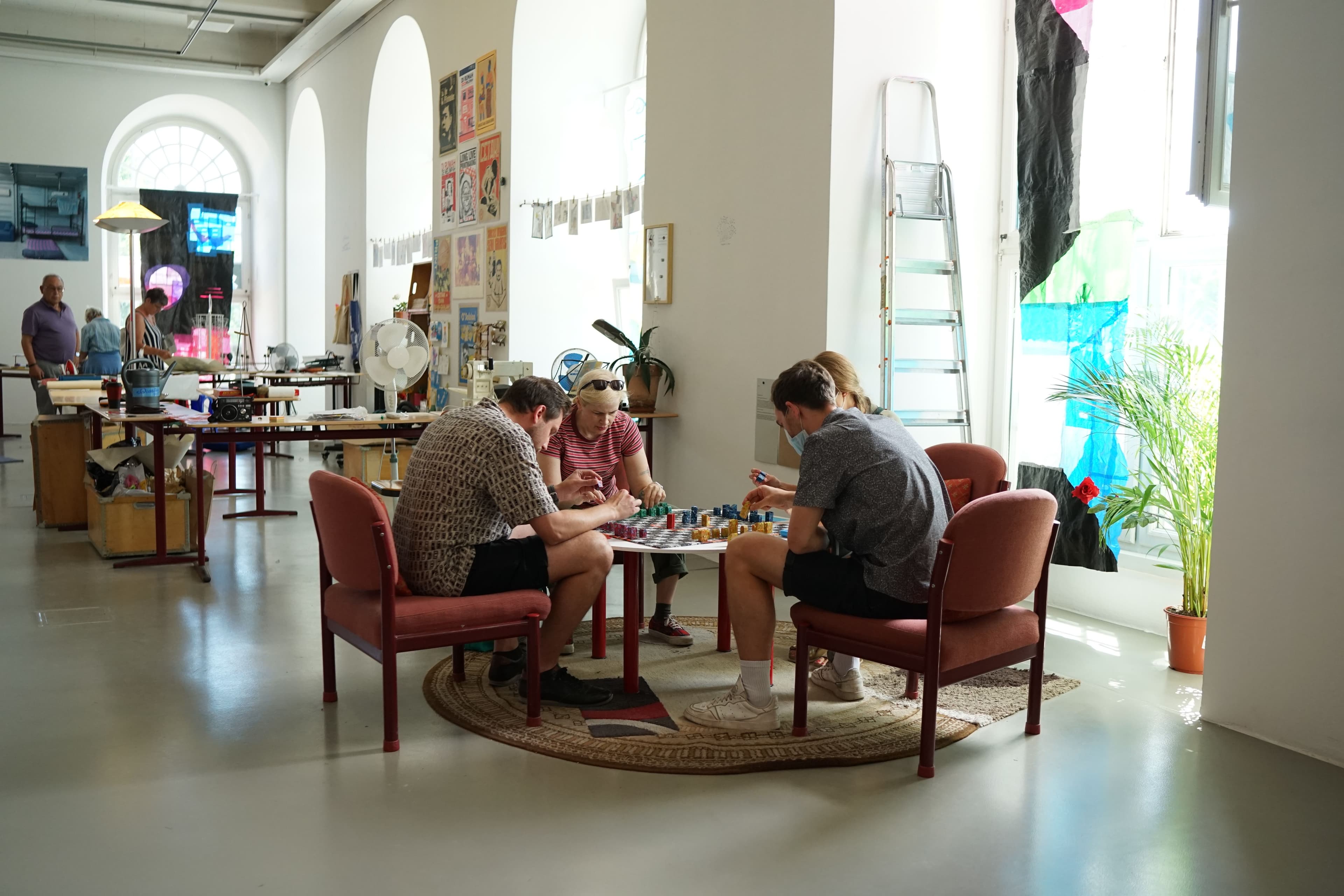 Four people sitting on low chairs around a small round table. They are playing a game of colorful bars on a checkered board.