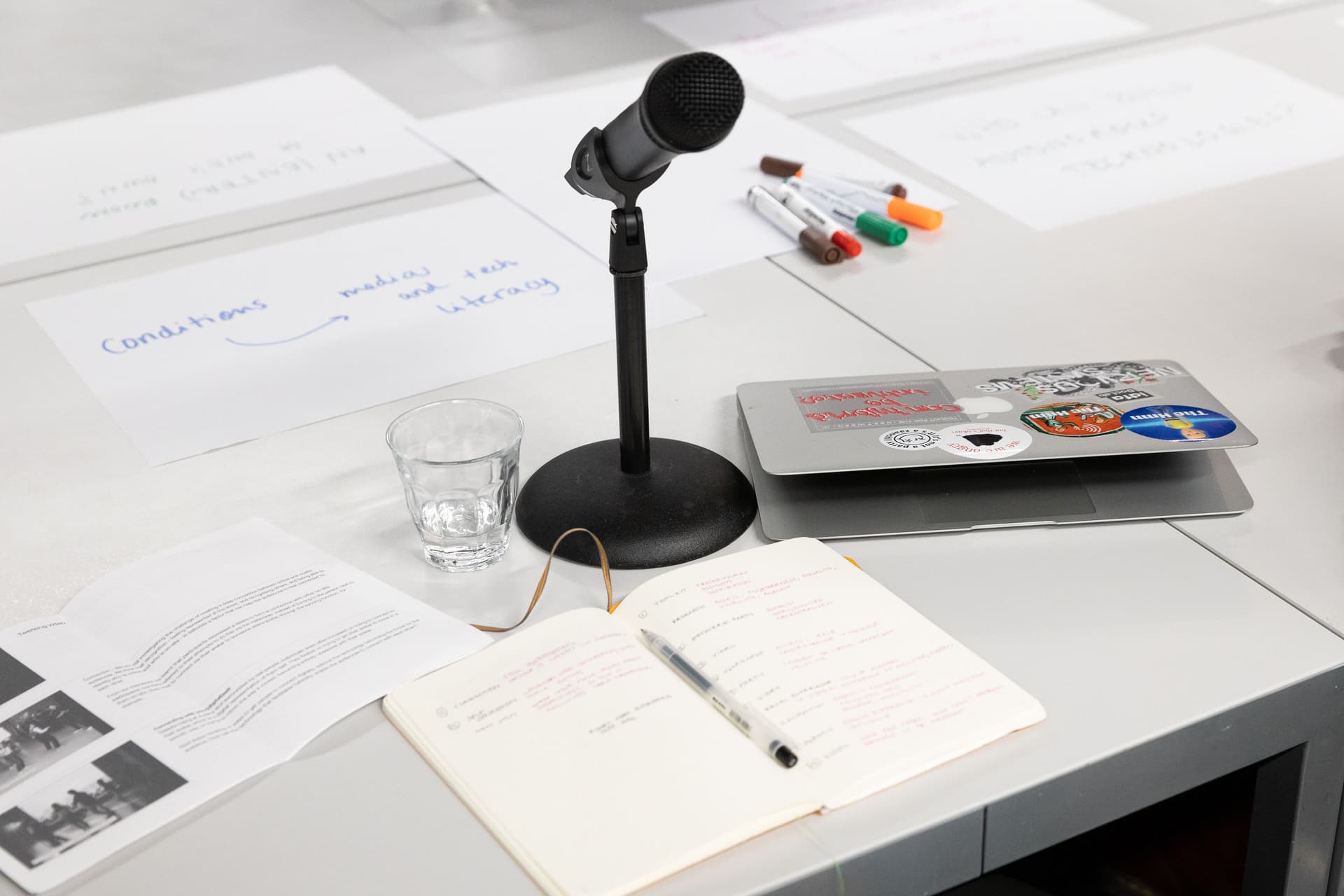  Messy white table with written-on paper, coloured markers, a microphone, glass of water and laptop
