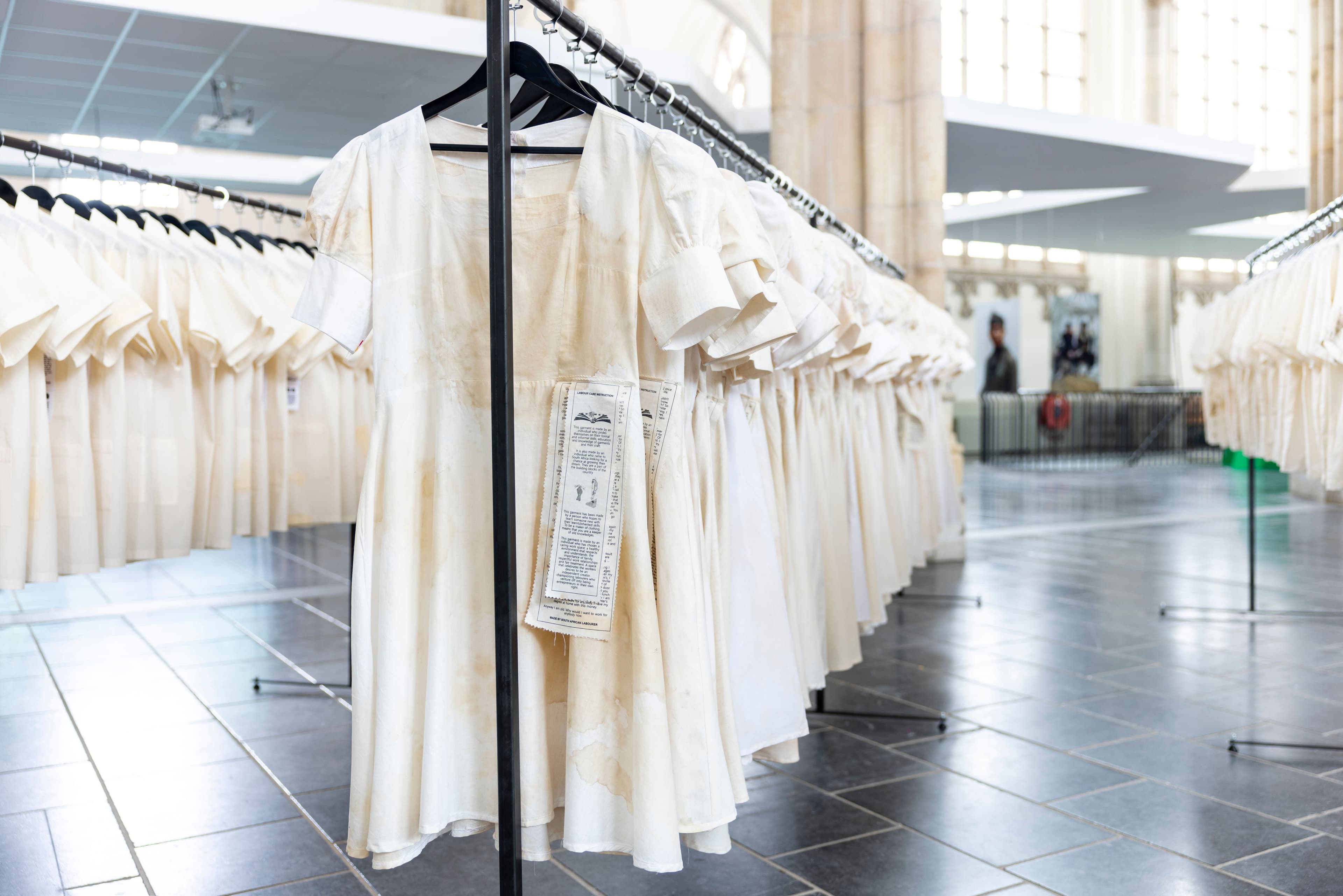 In an open space, filled with pillars and in the background stained glass, there are three rows of white clothing hanging from black clothing racks. The left and right rows have t-shirts. The middle row has dresses.