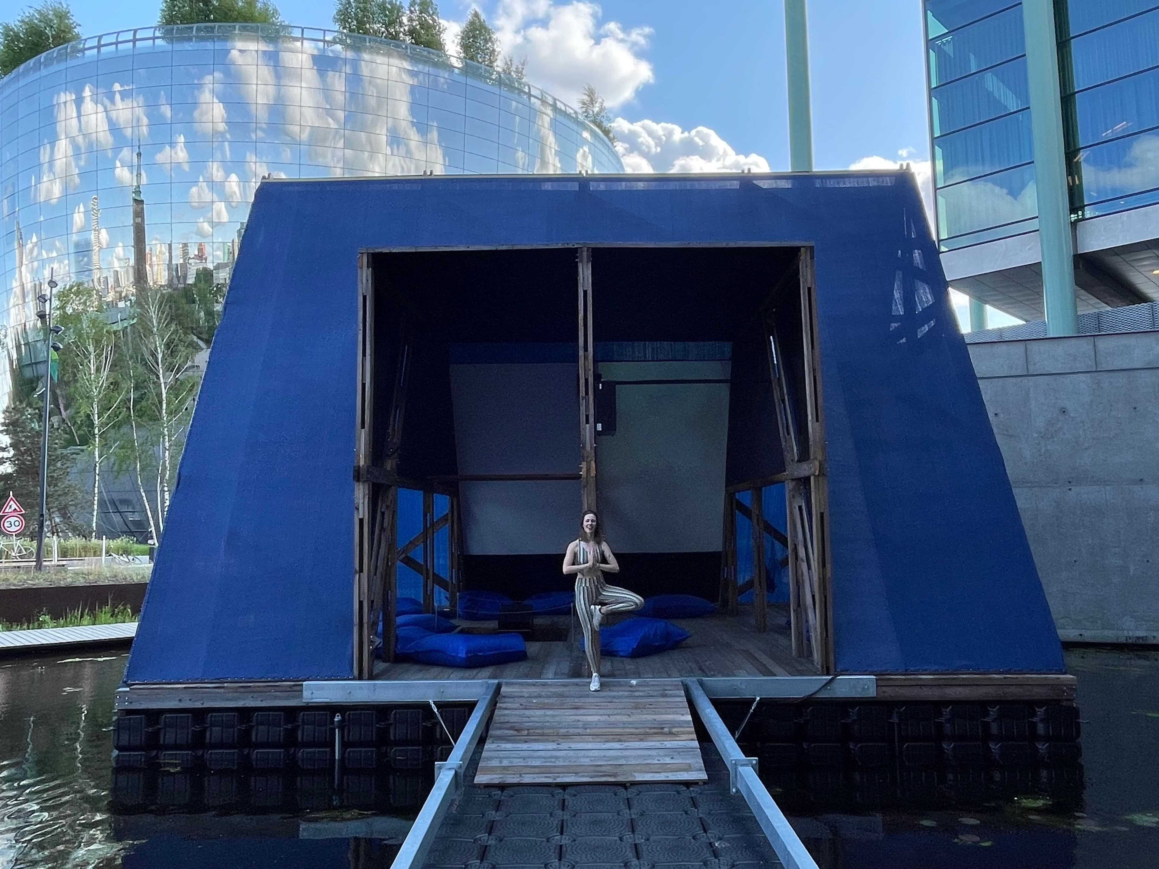 A woman is doing a yoga pose with hands in prayer position and one leg folded like a crane in front of the pavilion floating on the water