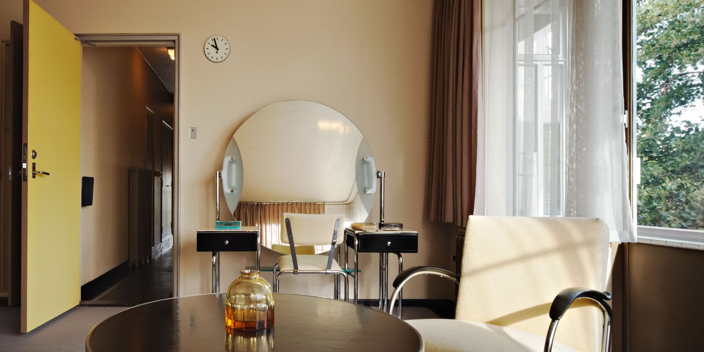 Een lichte kamer met moderne meubels, een ronde spiegel, een gele deur en een klok aan de muur.