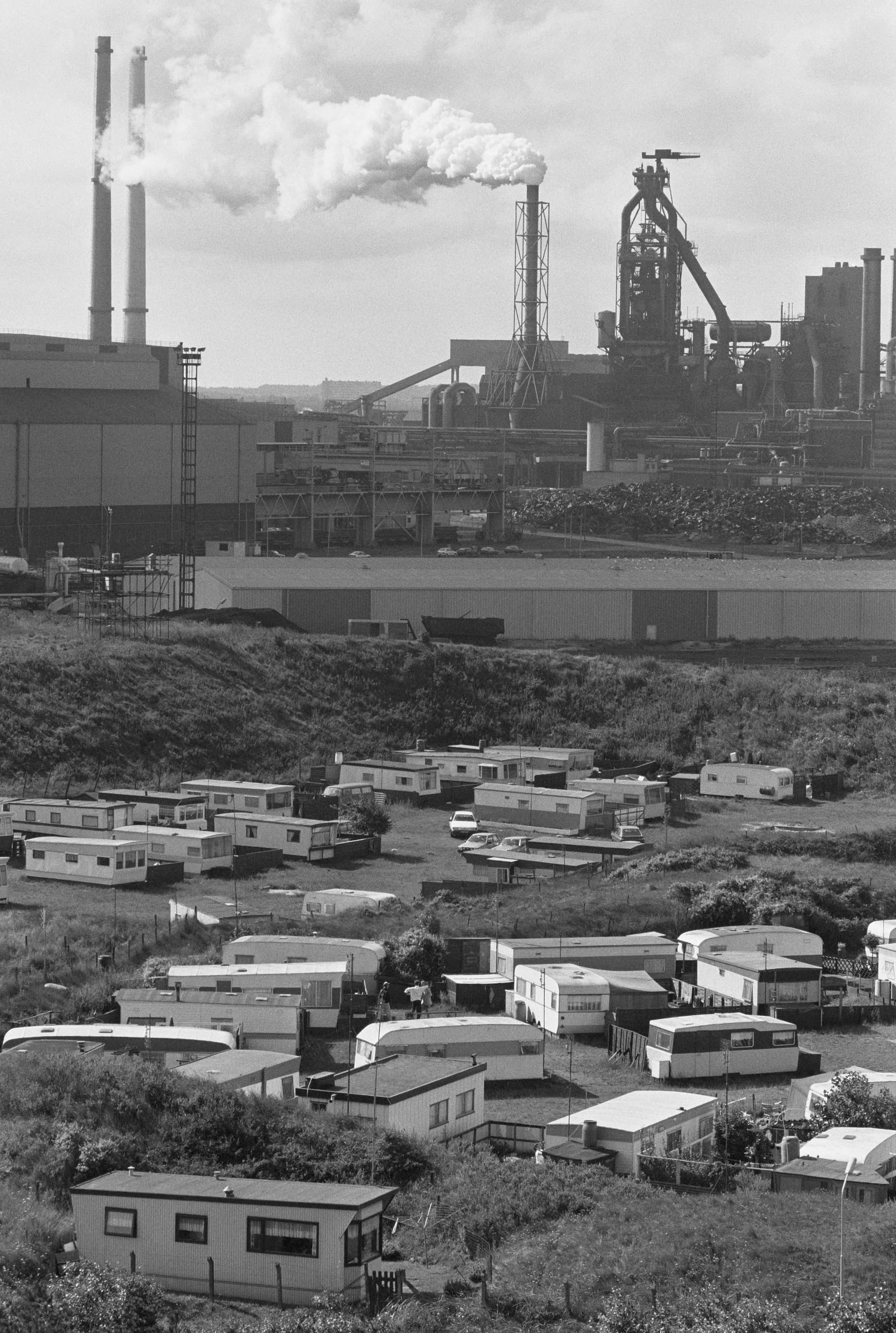 a contrast between a busy beach with beachcabins and people enjoying the sun versus polluting factories and industry further back.