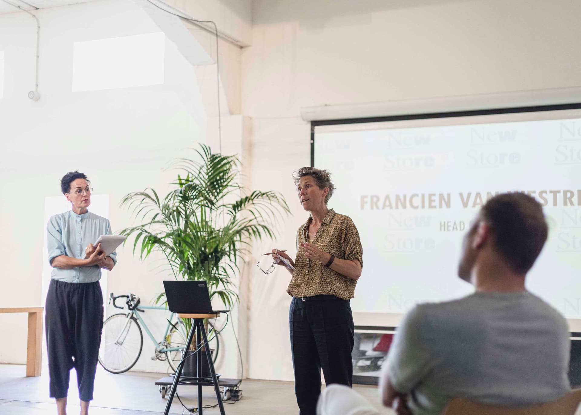 Een foto van de New Store-discussie. Twee sprekers staan voor een presentatiedia.