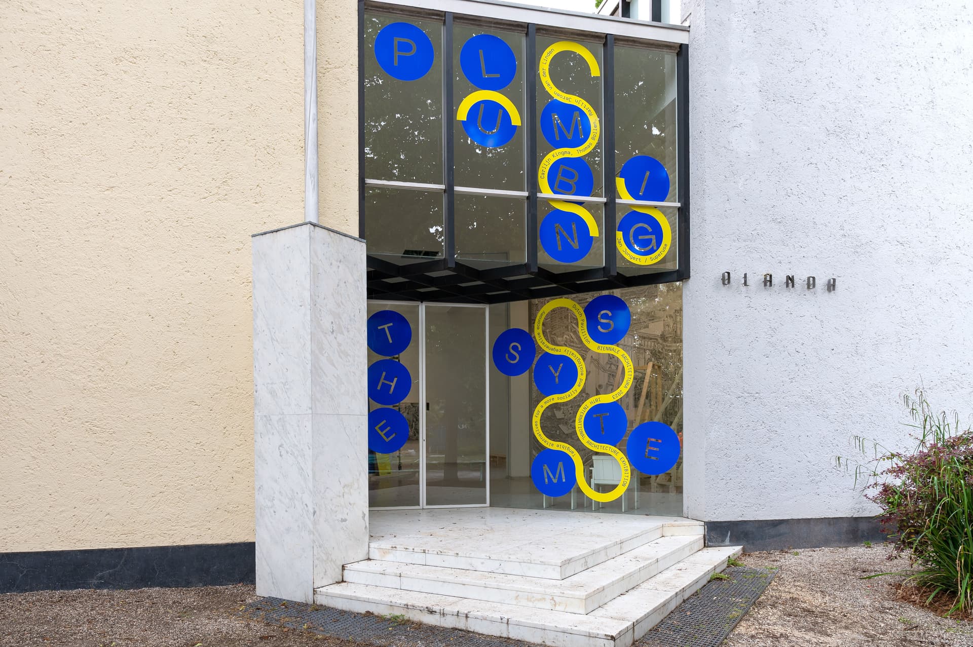 Entrance to the Rietveld Pavilion. shown is a glass door with a small staircase in front of it, a partly white and partly cream-coloured facade, and 'Olanda' on the wall.