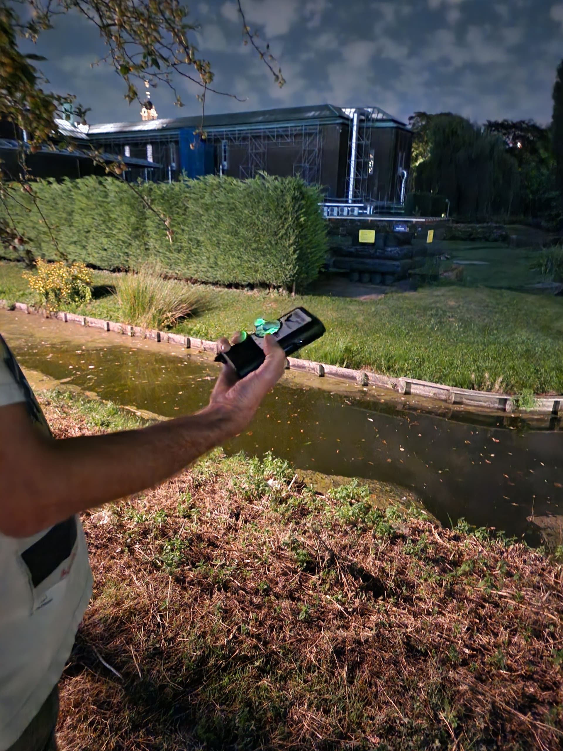 An outstretched arm holding a bat detector in the New Garden next to a ditch, at night.