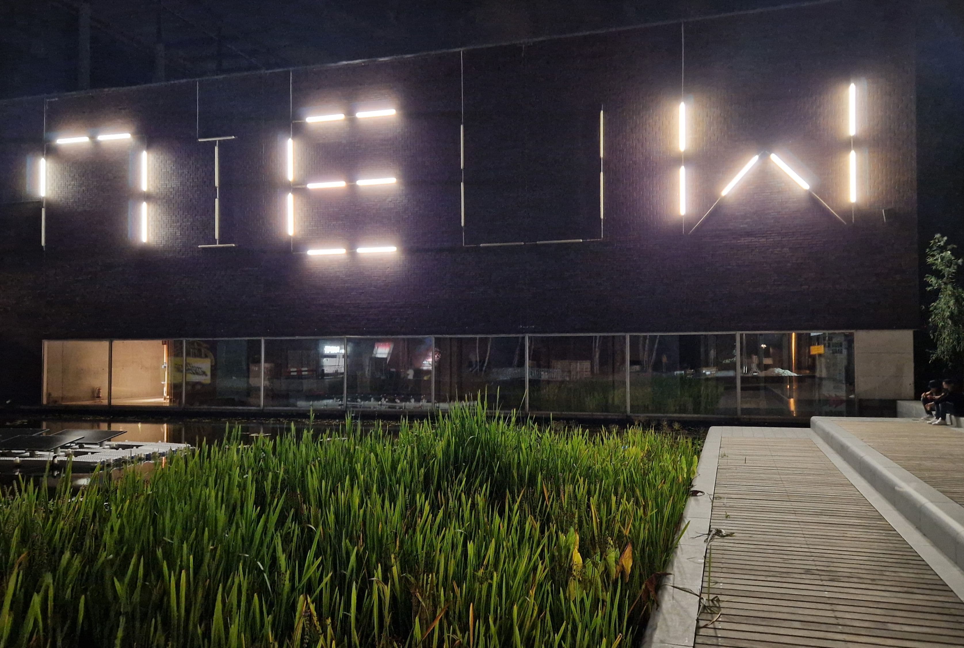 A view of Het Nieuwe Instituut at night, with the word 'NIEUW' in large, glowing letters on the outside, with the pond and reeds in the foreground.