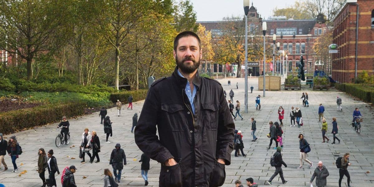 A man, Arne Hendriks, is standing in a square of gray tiles. In the background on the left is a park with low greenery and taller trees, buildings on the right and lampposts in the middle. The man is surrounded by walking and talking passers-by… 
