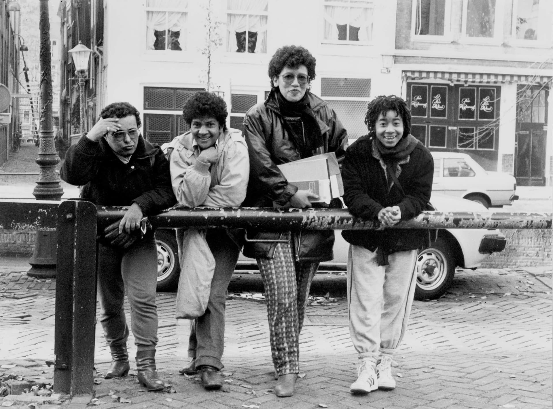 Foto in zwart-wit van vier vrouwen naast elkaar, leunend tegen een hek. Op de achtergrond staat een auto en op de achtergrond zijn verschillende stadspanden. 