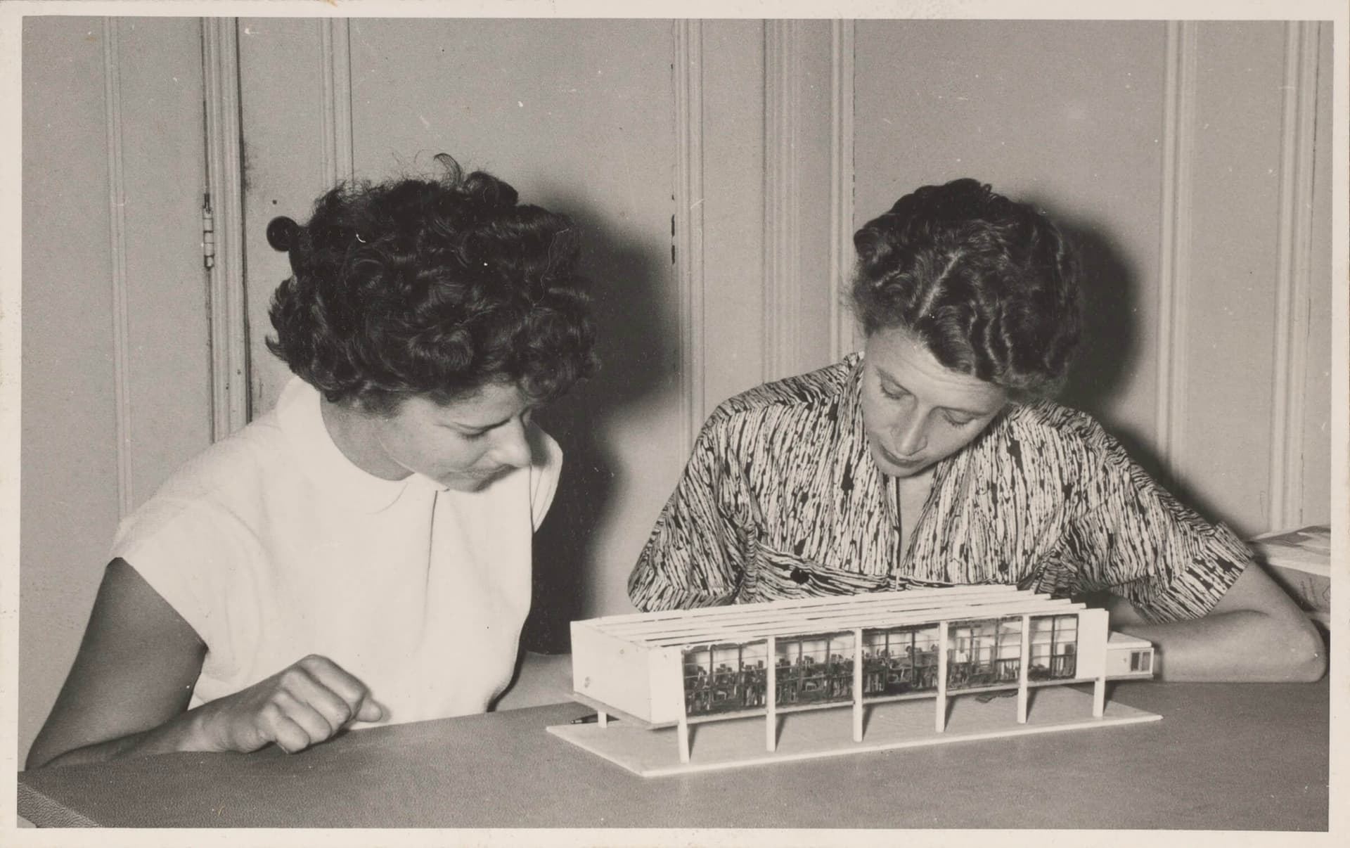 Zwartwitfoto. Twee vrouwen bestuderen een architecturaal model van een gebouw.