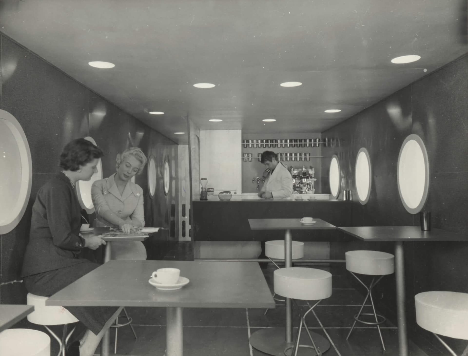 Zwartwitfoto van een treincoupé met tafeltjes, ronde ramen en een bar.