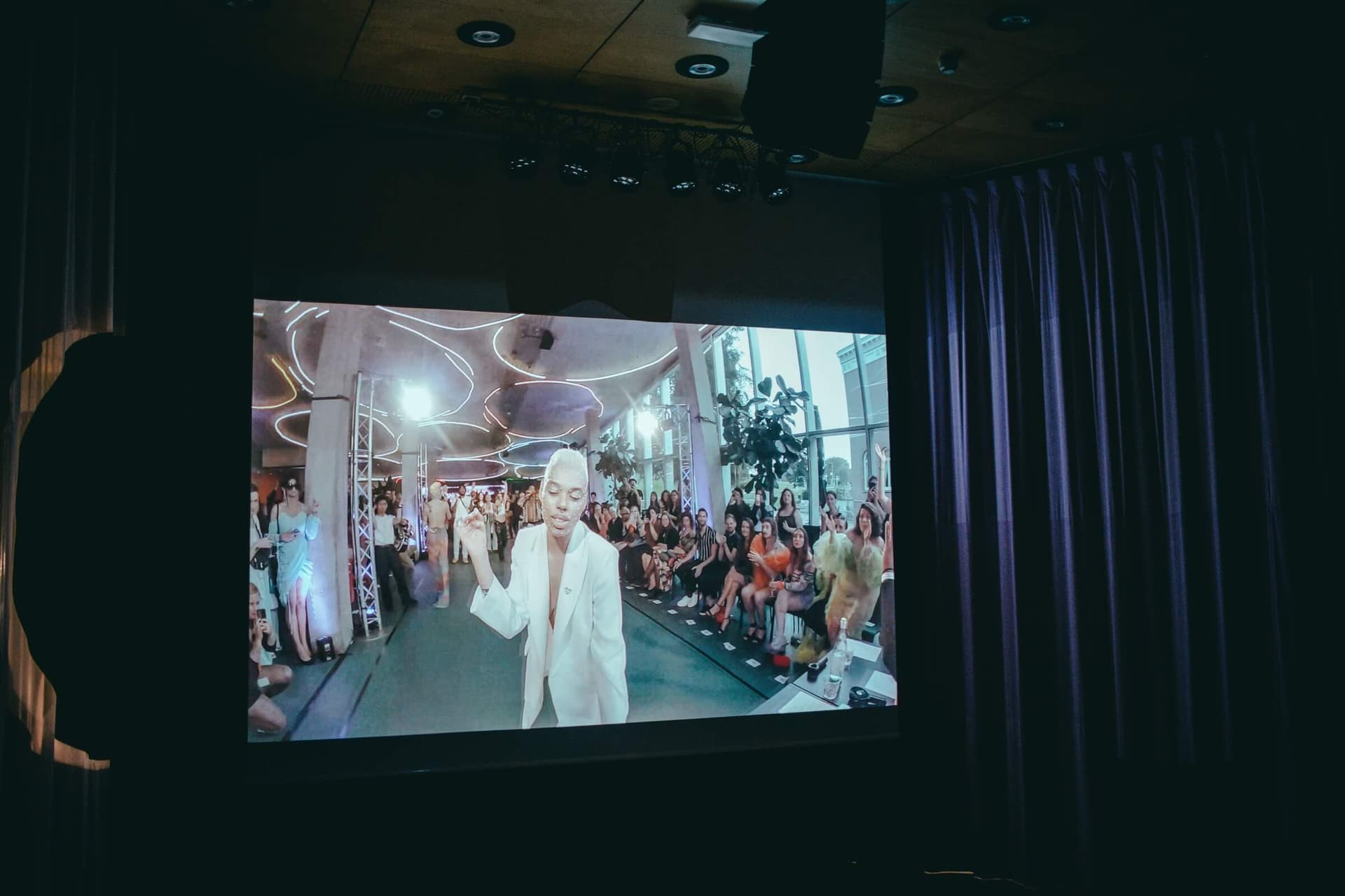 Photo of a dark room with a screen. The screen shows a person wearing a white blazer looking towards the camera with his eyes closed. This person is standing on a green floor with people on two sides; it looks like a catwalk, made in the K… 