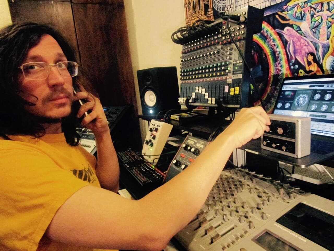 Danny wolfers aka Legowelt. Op de foto staat een man met lang donker haar en een bril. Hij draagt een geel t-shirt. Hij zit in een muziekstudio, achter een bureau met daarop verschillende synthesizers, speakers en andere audioapparatuur. 