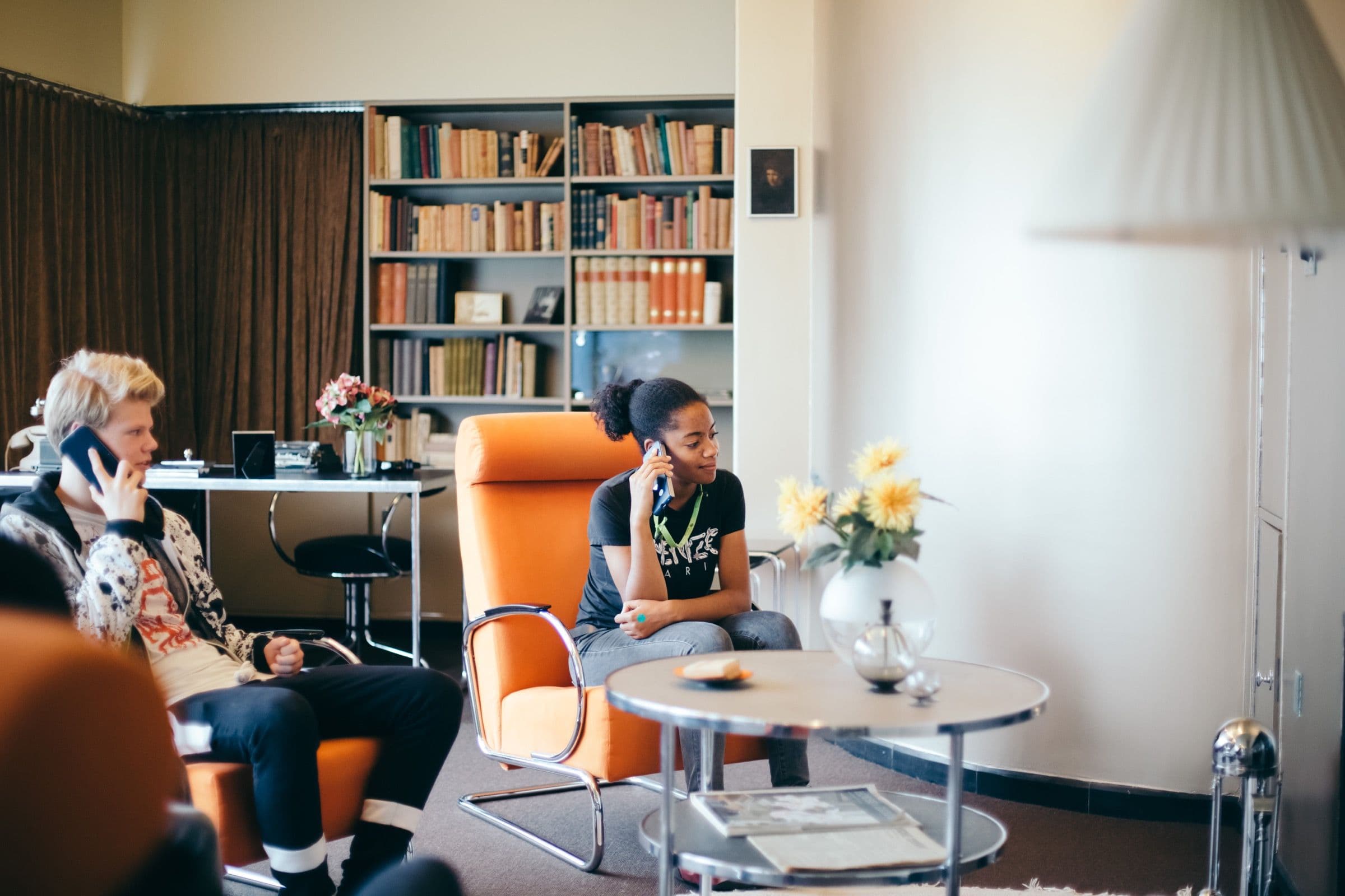 Twee mensen zitten in oranje stoelen en bellen, omringd door een retro-interieur met boekenplanken, een tafel met gele bloemen en een rustige, stijlvolle sfeer.