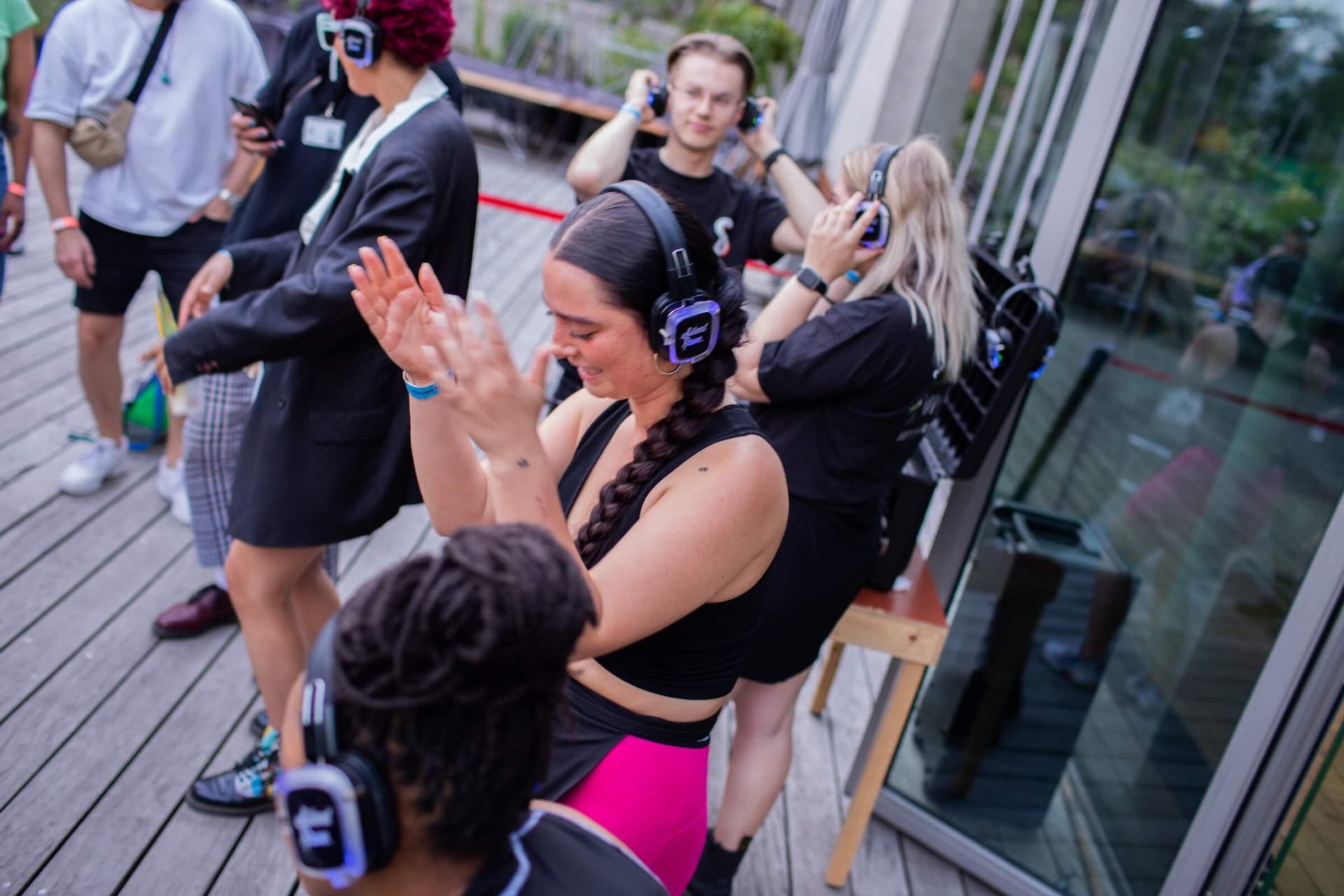 A group of young people wearing headphones dance on the terrace of Het Nieuwe Instituut The headphones have a blue light. In the middle, a young woman with black hair and a long braid claps her hands.   