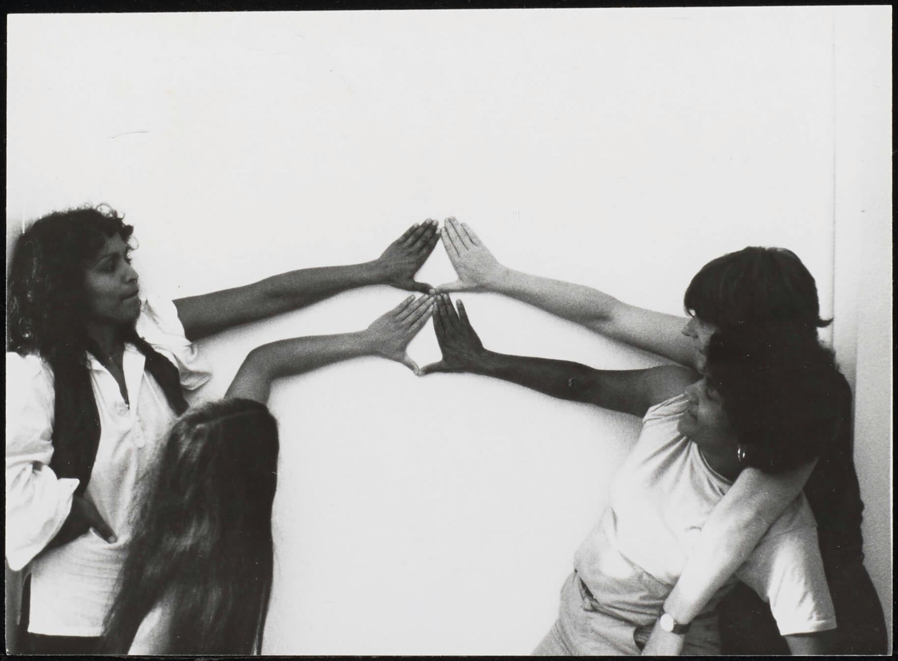 Vier vrouwen vormen driehoeken of het Yoni-teken met hun handen tegen een witte muur: tegen de klok in vanaf rechtsonder: Tania Leon, Lida van den Broek, Ananda Spies, Margrethe Rumeser, jaar: 198?, foto: Gon Buurman. Bron: Collectie IAV-Atria   