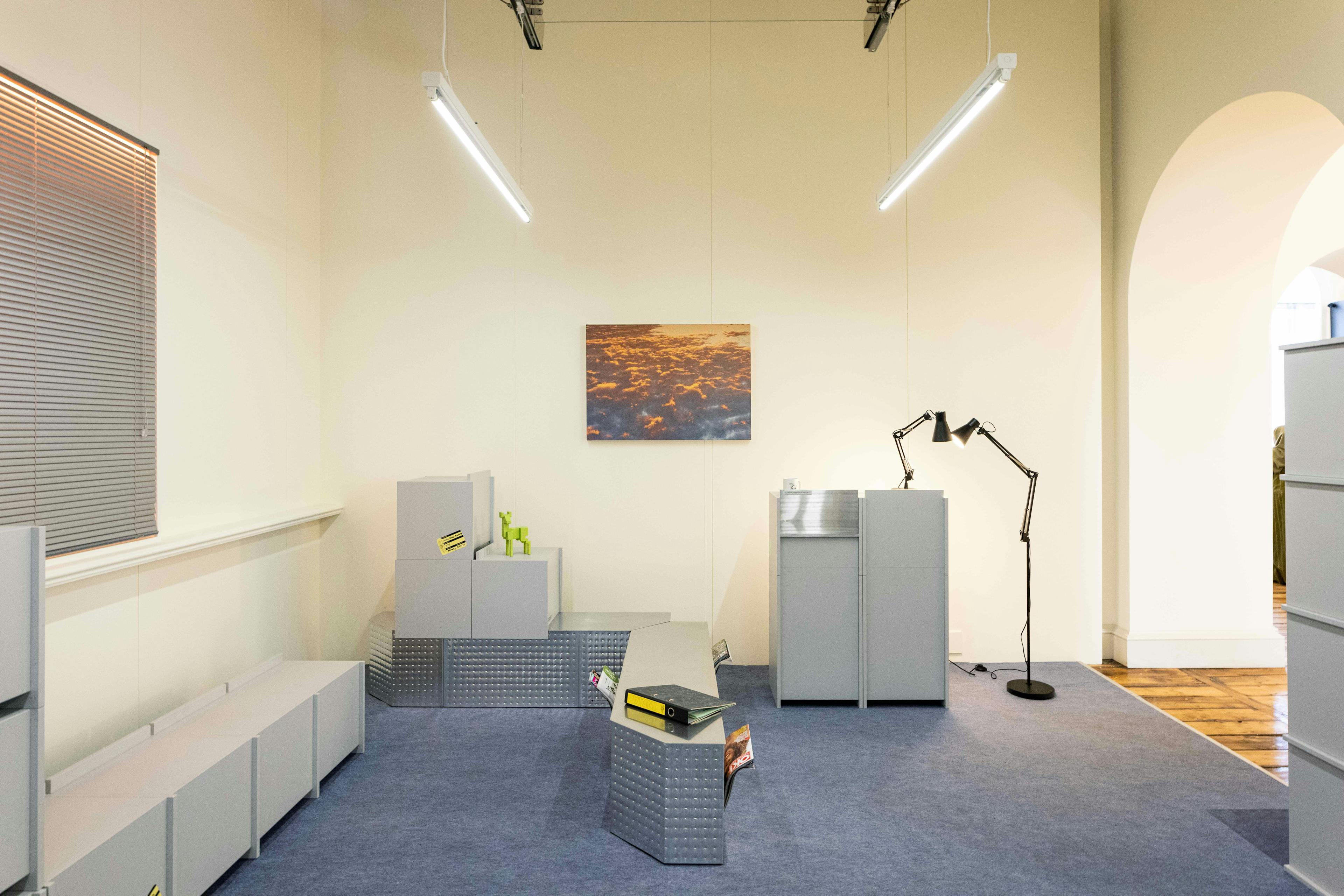 Een tentoonstellingsruimte met objecten die stereotiep een kantooromgeving voorstellen: kasten, lampen, TL-verlichting, etc.