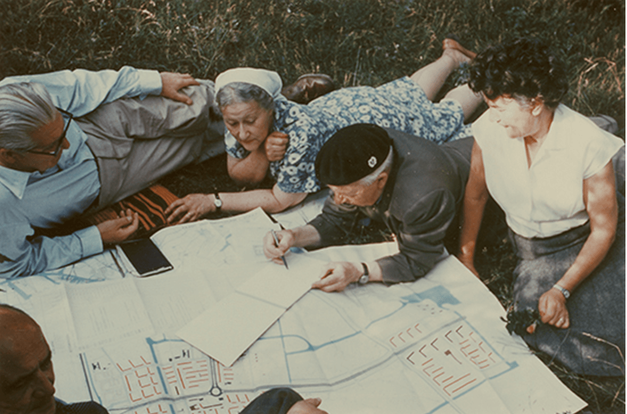 An archival image displaying a group of older people, lying on the grass in a circle around a large map. One older man in the center is jotting something down in a notebook as the two women, each on one side, watch.
