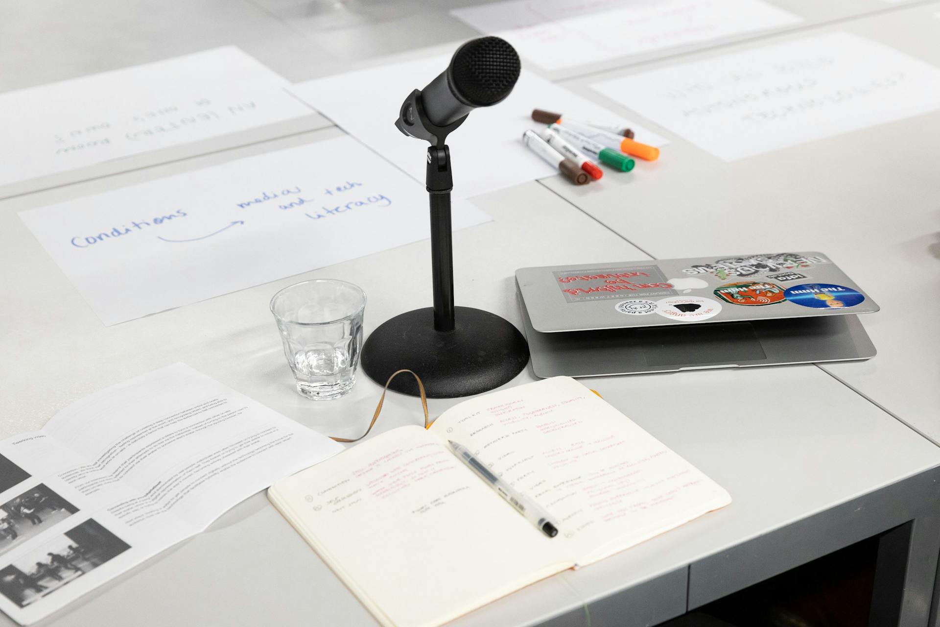  Rommelige witte tafel met beschreven papier, gekleurde markers, een microfoon, glas water en laptop