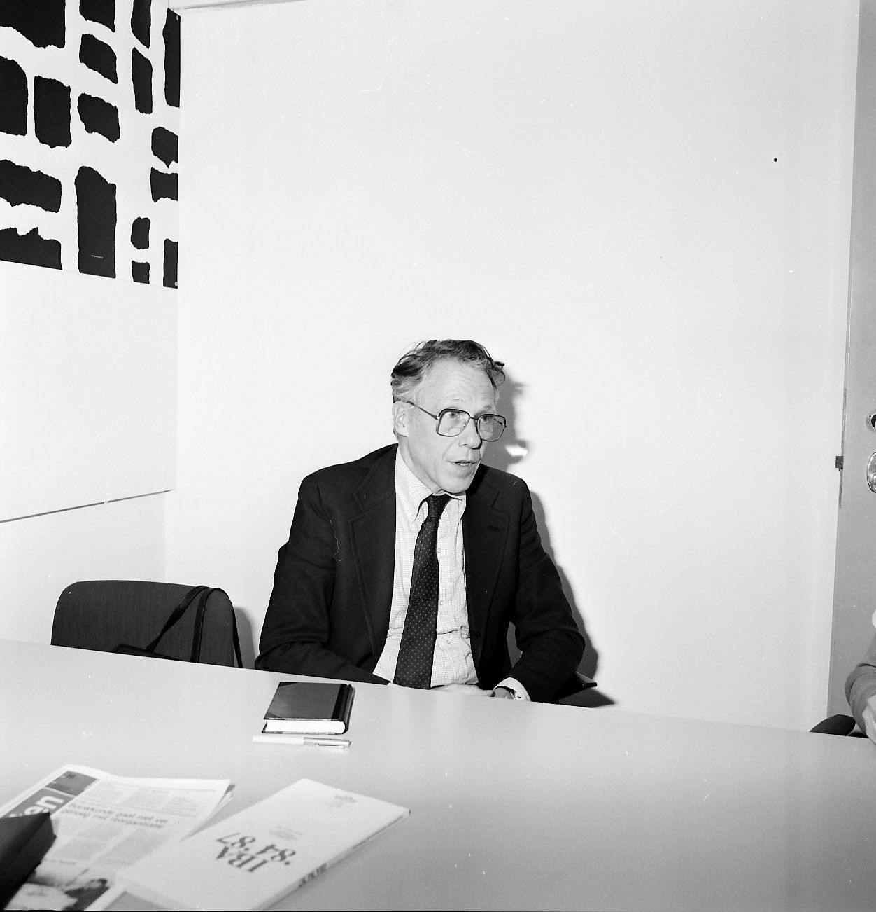Portret van John Habraken in zwart/wit, zittend achter een bureau in een werkkamer, mogelijk op de Universiteit van Eindhoven.