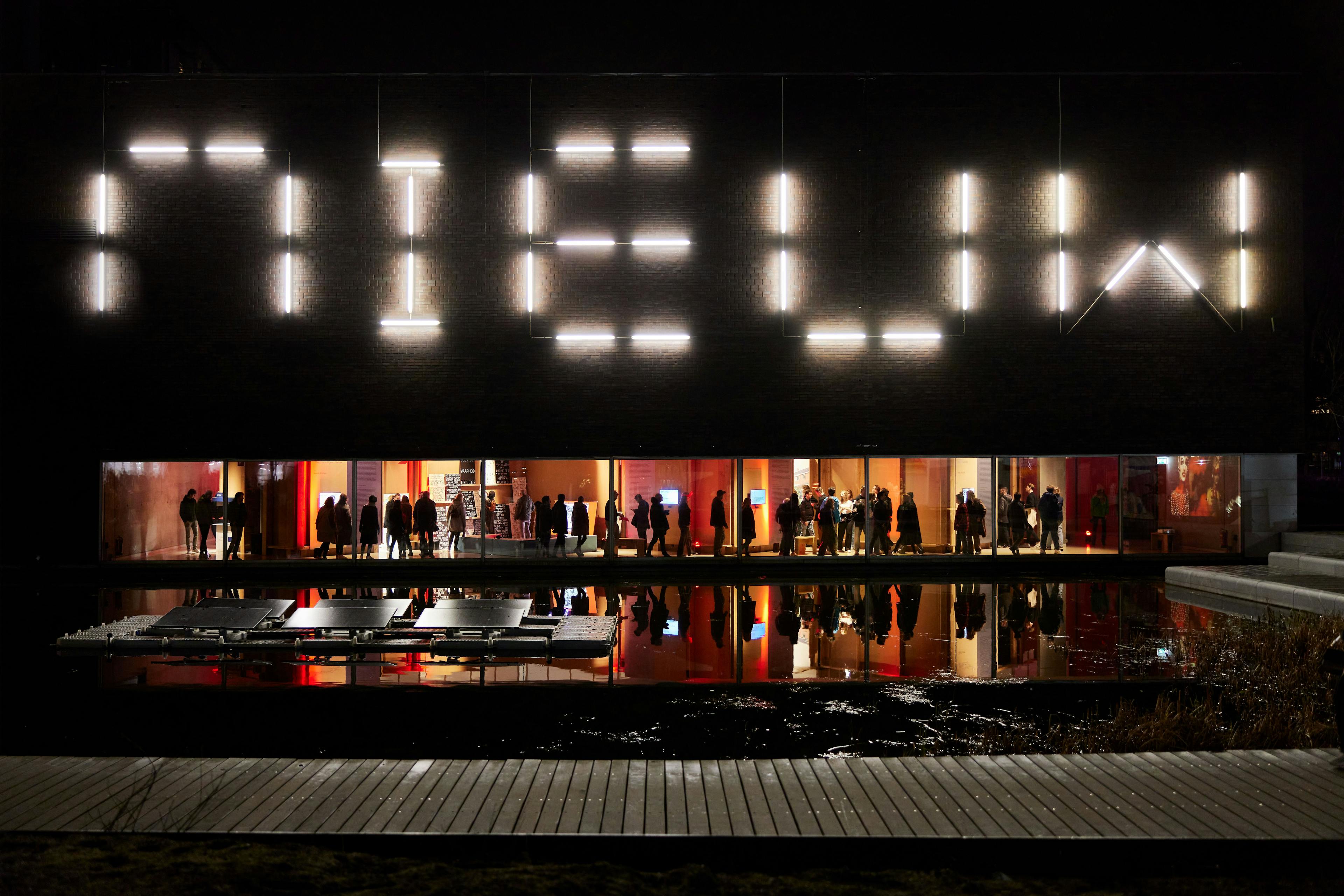 In the dark the letters NIEUW light up, in the centre a long stretched window with red light and people.