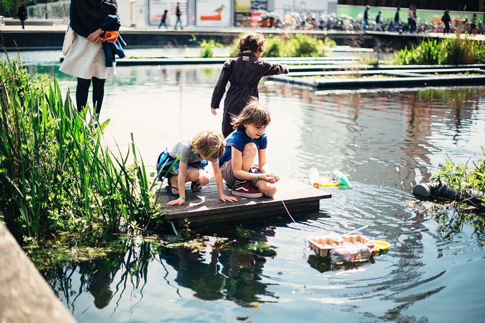 On a wooden platform, near the water around the institute, two kids are squatting. One of them is holding a rope with a small float tied to it made out of cardboard materials.