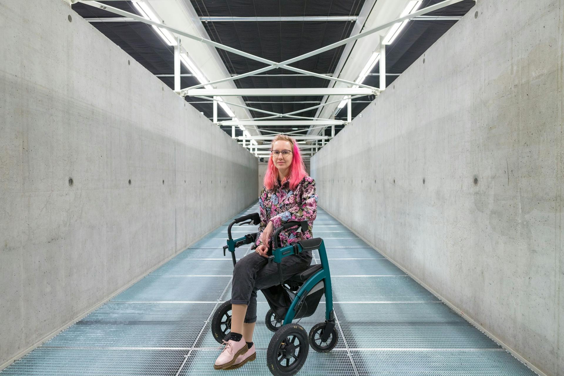 Picture of a pink-haired woman in a wheelchair in the middle of an industrial looking hallway.