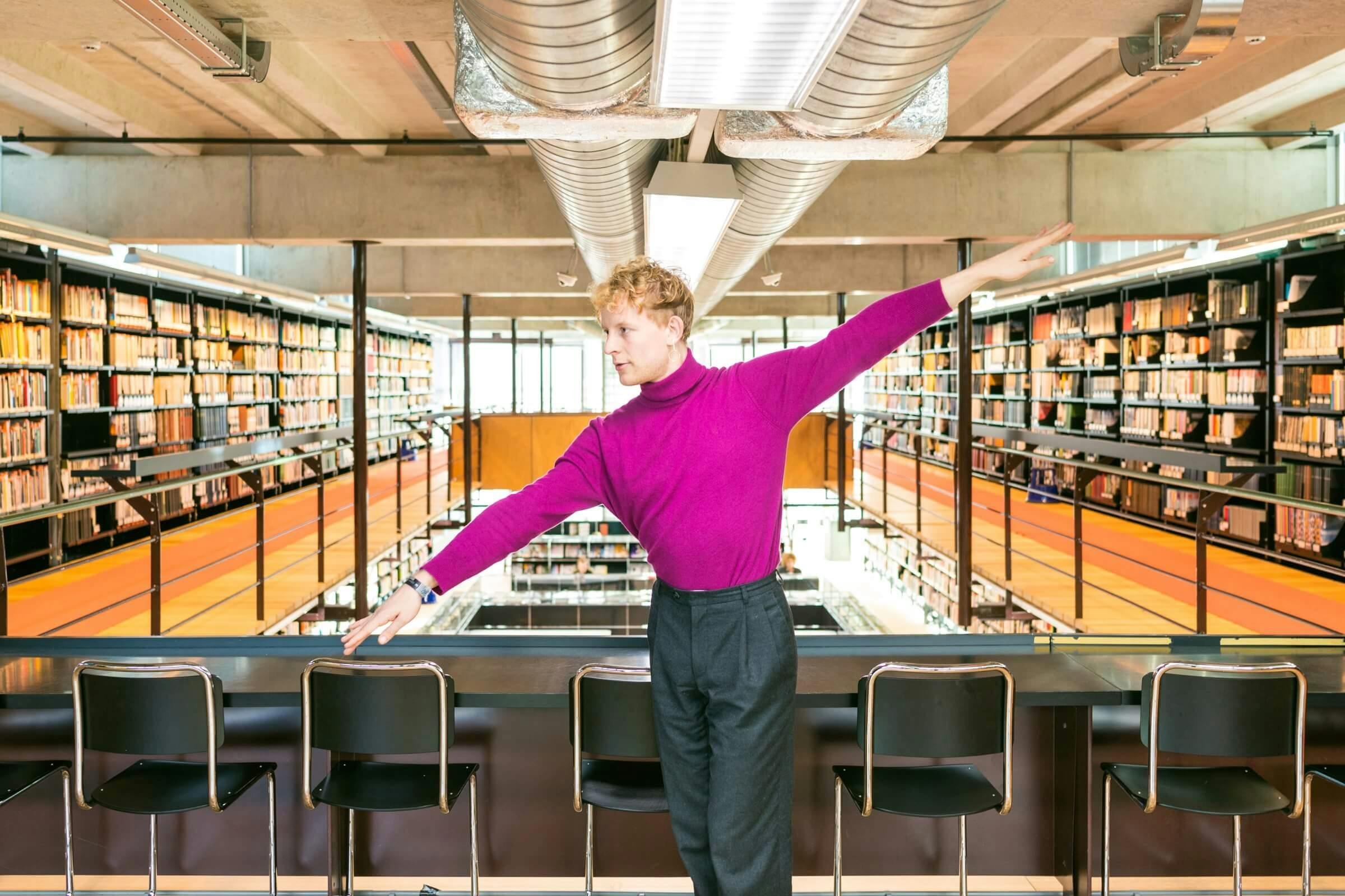A man in bright pink top and grey trousers, spreading his arms to opposite sides in a library